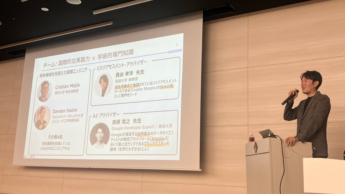 国際的な実装力と学術的知識を組み合わせたチーム紹介スライドを投影しながら発表する様子。リスクアセスメントやAIアドバイザー陣のプロフィールが表示されている。